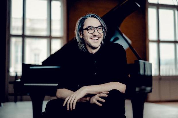 Uomo con occhiali e capelli lunghi brizzolati seduto. Dietro di lui, un pianoforte.