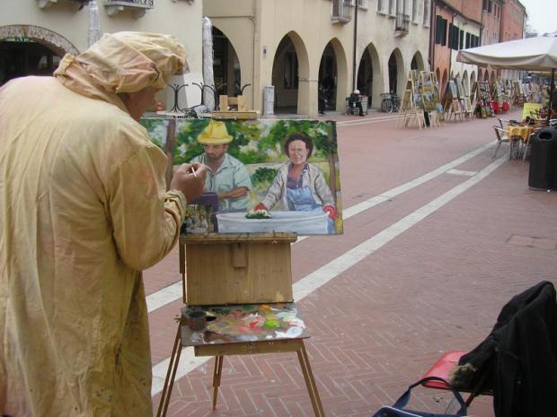 Pittore, visto di spalle, lavora al suo quadro, posto su un cavalletto, all'aperto, in una via cittadina