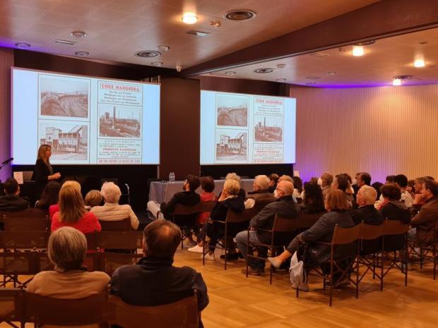 Pubblico seduto assiste a una conferenza nella quale una donna parla di fronte a due lavagne luminose