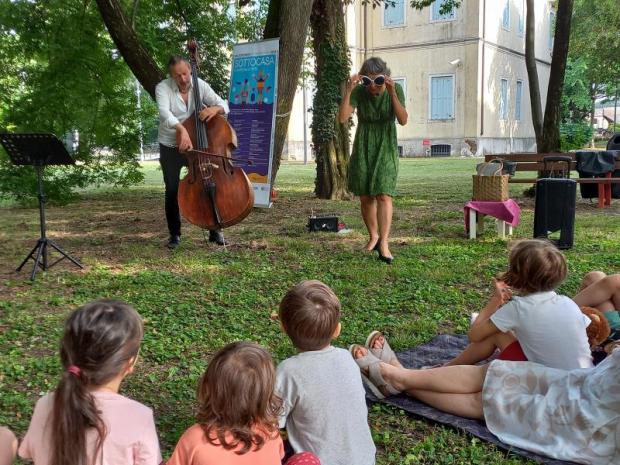 Davanti a pubblico di bambini (di spalle) si esibiscono un'attrice con occhiali scuri e un contrabbassista