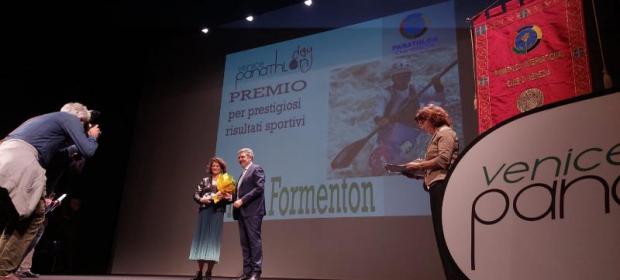 Una donna, con un mazzo dei fiori in mano, viene premiata da un uomo sul palco. In primo piano, a sx, un fotografo