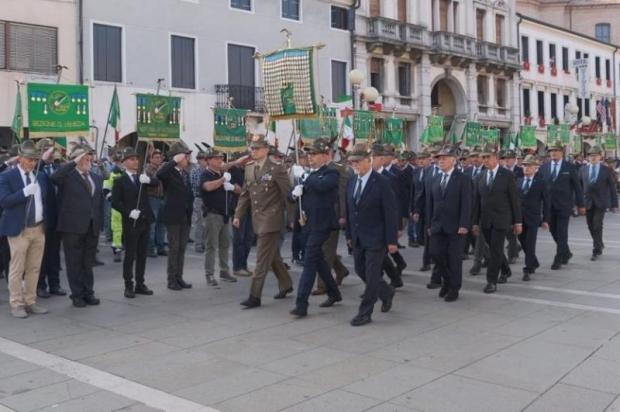 Gruppo di uomini anziani, in abito o divisa, col cappello da alpino sfilano in una via cittadina reggendo degli stendardi