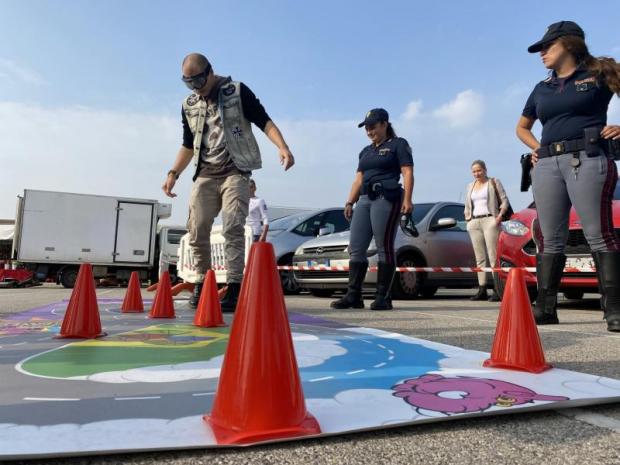 Due poliziotte guardano un uomo bendato che affronta un percorso-gioco allestito con dei coni per il traffico