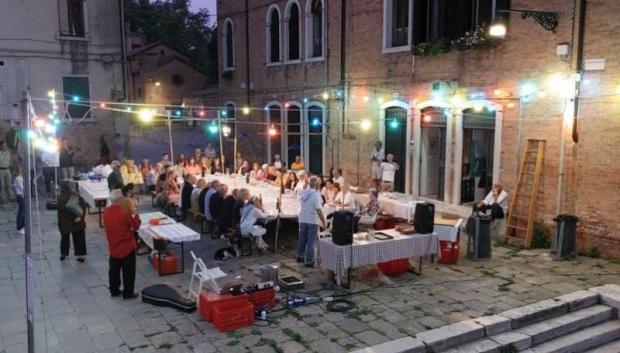 Decine di persona a una grande tavolata allestita all'aperto di sera in un campo di Venezia