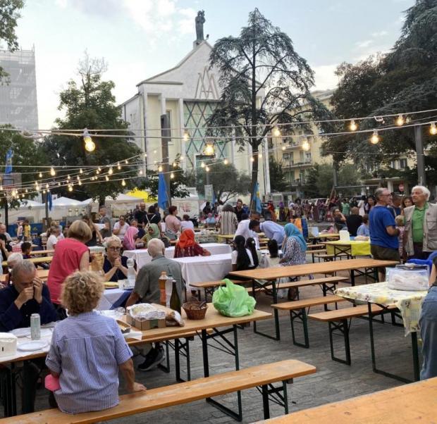 Persone sedute a tavoli da sagra sotto filari di lampadine. Sullo sfondo, una chiesa