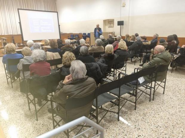 Pubblico, visto da dietro, segue una conferenza. In fondo il relatore parla accanto a uno schermo