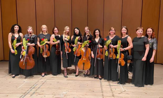 Foto di gruppo di 12 donne disposte in riga, in piedi. 9 di loro tengono in mano uno strumento musicale a corde