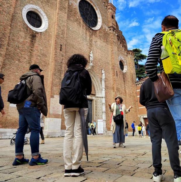 Gruppo dii persone viste di spalle che ascolta una guida, di fronte, davanti a facciata in mattoni di chiesa veneziana