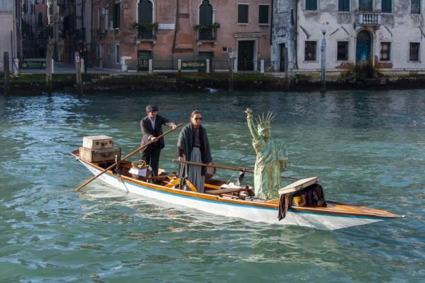 Due persone in maschera vogano su una barca in cui è caricata una riproduzione della Statua della Libertà