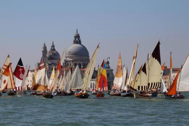 Decine di imbarcazioni dalla vela colorata navigano sulla laguna di Venezia. Sullo sfondo, una chiesa
