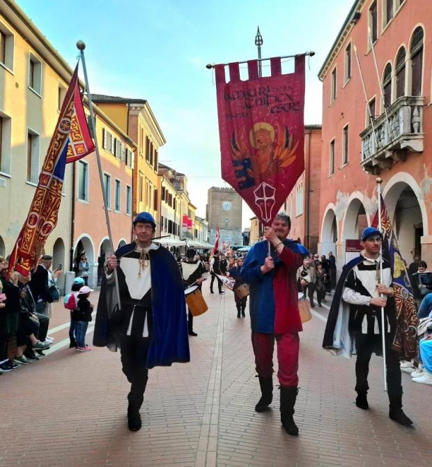 Sfilata di figuranti in abiti medievali tra due ali di pubblico. Sullo sfondo una torre con orologio