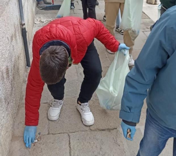 Ragazzino si piega in avanti per raccogliere da terra un mozzicone e metterlo nel sacco di rifiuti che tiene in mano