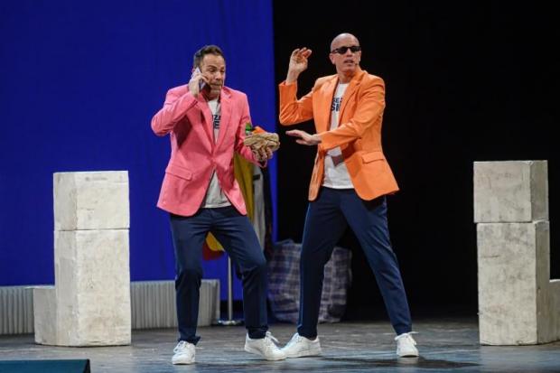 Due comici (Carlo&Giorgio) sul palco, vestiti con giacca e pantaloni. Quello a sx regge il cell, quello a dx ha occhiali da sole