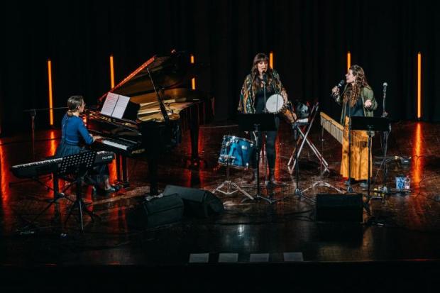 Tre musiciste sul palco in concerto: una al piano, una ai tamburi, una al microfono